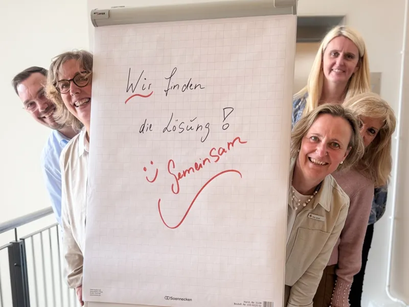Ein Teil des Teams der Betriebs- und Prozessorganisation steht um einen Flipchart auf dem geschrieben steht: Wir finden die Lösung gemeinsam!